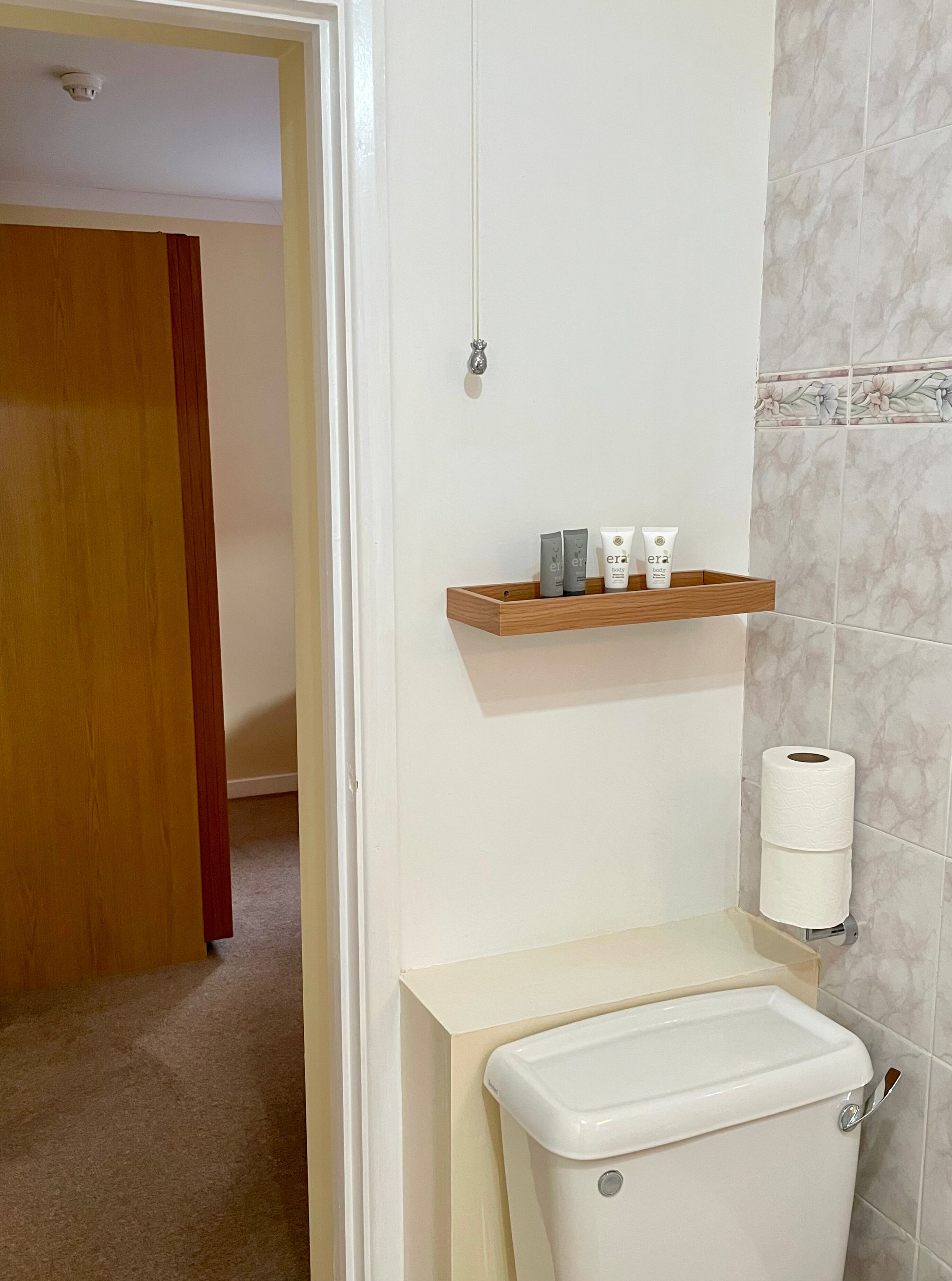 A bathroom with a toilet, a wooden shelf with toiletries, and a toilet paper holder on the wall.