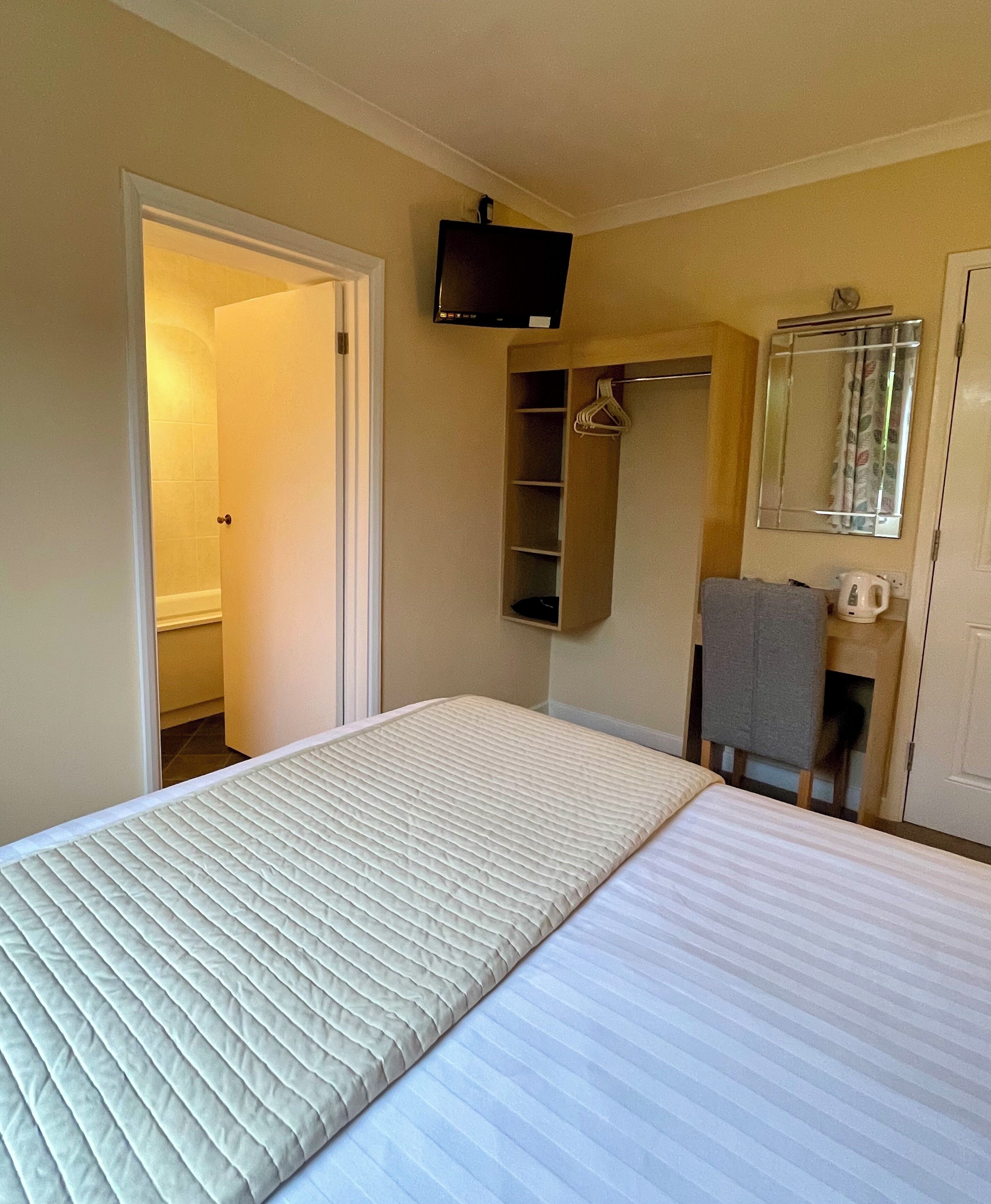 A hotel room with a bed, a television mounted on the wall, a mirror, a towel rack, and a small shelf.