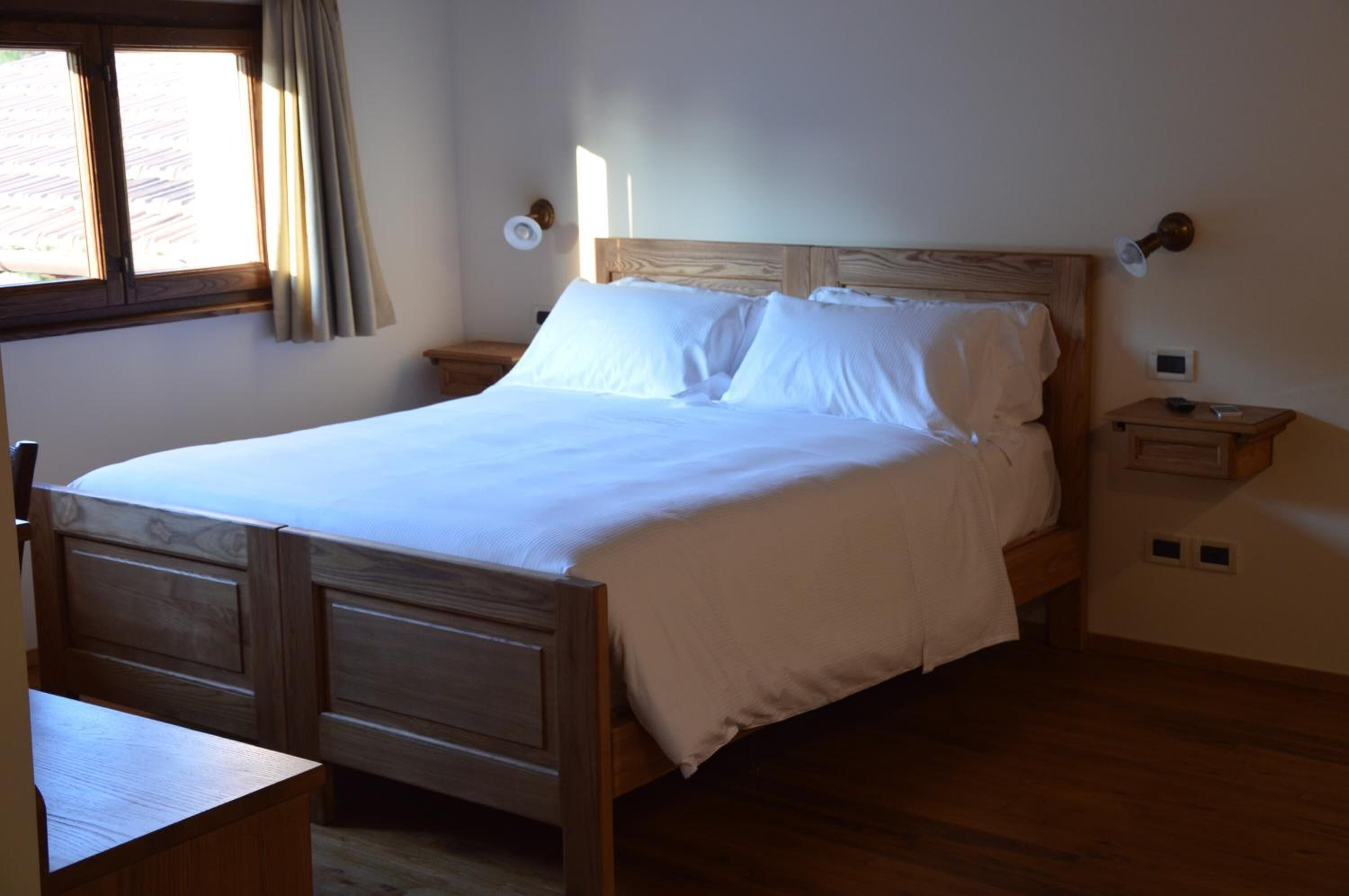 A wooden bed with white linens in a room with a wooden floor and a window with curtains.
