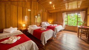 Habitación con paneles de madera, con tres camas, cada una con colcha roja y ropa de cama blanca. Hay mesitas de noche con lámparas, una silla de madera y una ventana con cortinas.