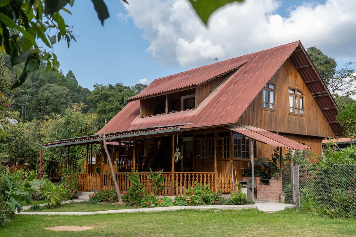 Een rustiek houten huis met een schuin dak, een veranda en een tuin met verschillende planten.