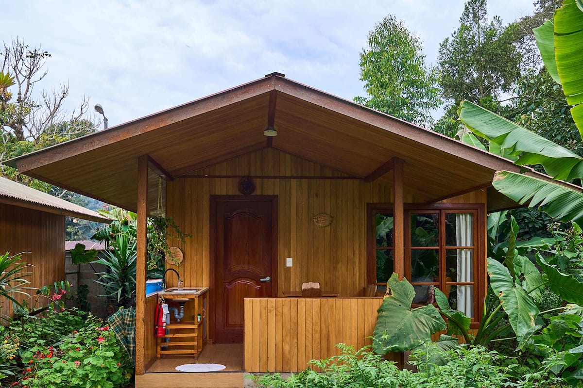 Een houten chalet met een veranda, omgeven door weelderige begroeiing en een betonnen pad dat naar de ingang leidt.