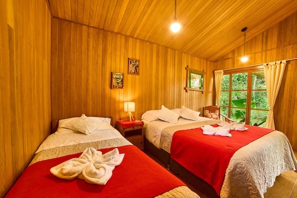 A wooden room with two beds, each with a red bedspread and white pillows. There is a nightstand with a lamp and a framed picture on the wall.