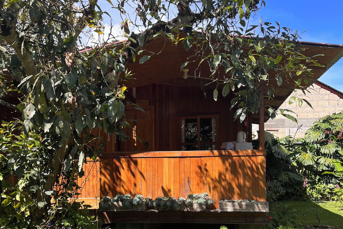Een houten hut met een veranda, omgeven door weelderige begroeiing en een heldere blauwe lucht.