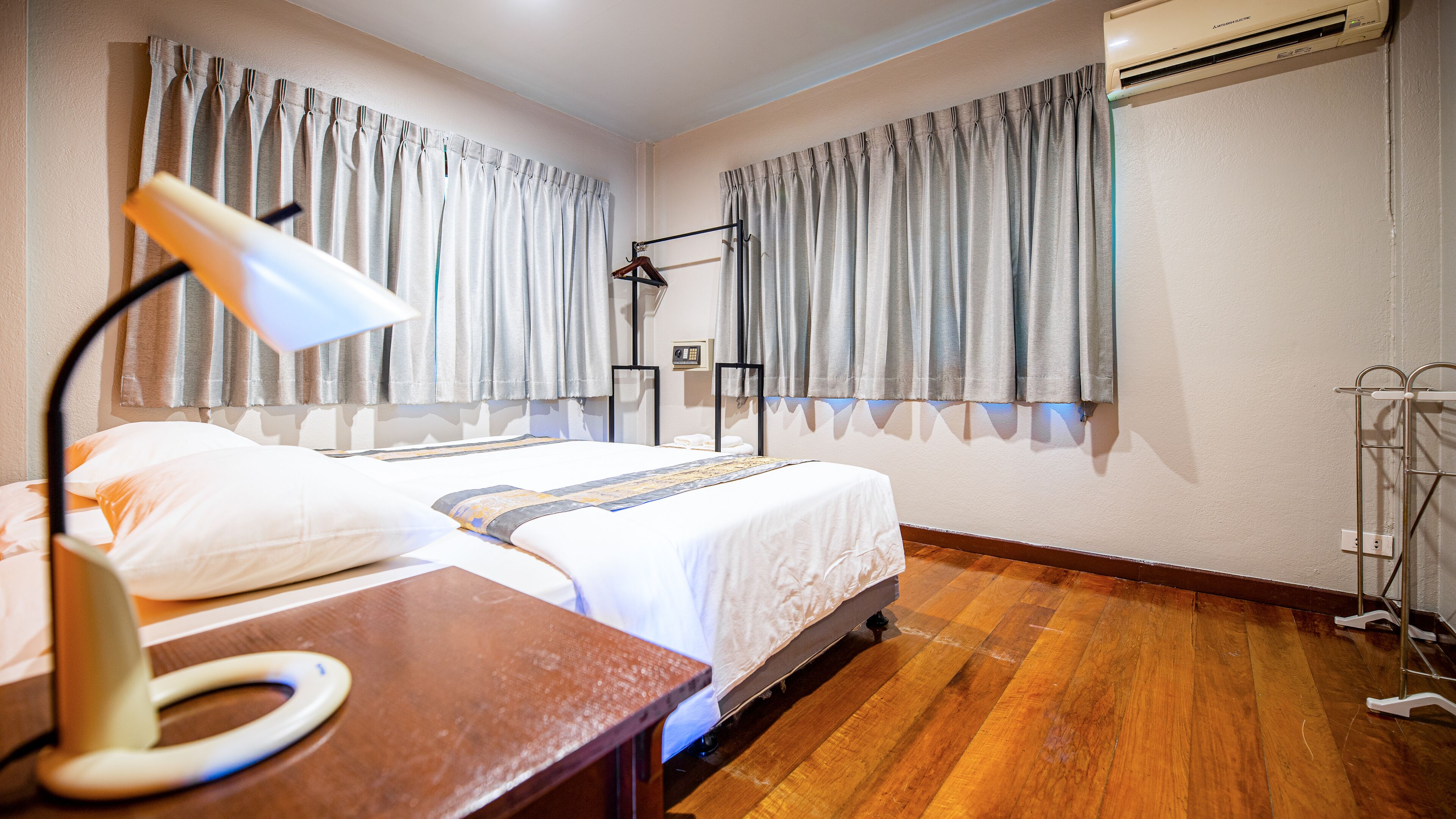 A hotel room with a bed, a desk with a lamp, a wardrobe, and an air conditioning unit.