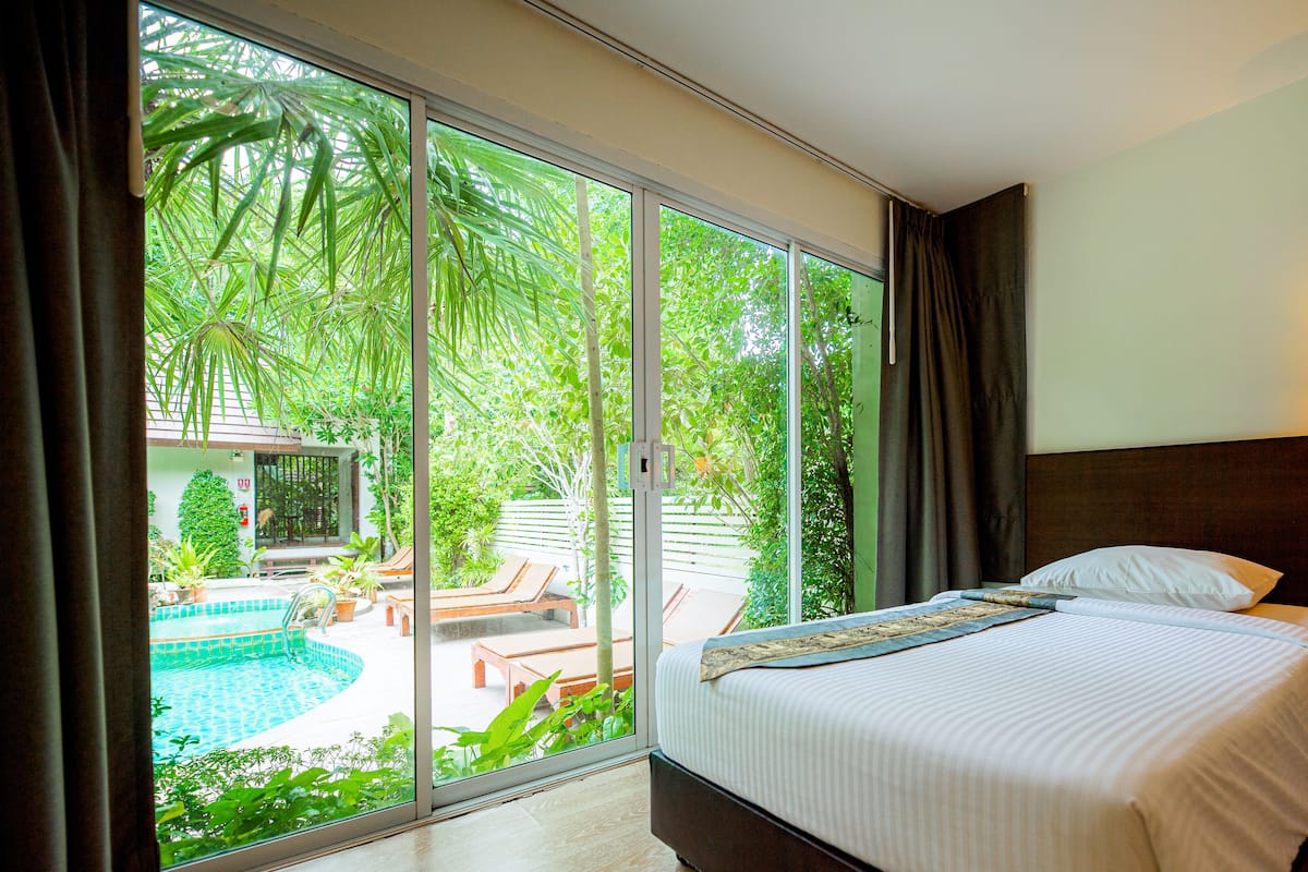 A hotel room with a bed, a nightstand, a lamp, and a view of a swimming pool and greenery outside.