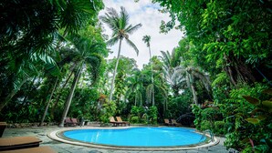 Una piscina rodeada de exuberante vegetación y palmeras, con sillones de descanso y un camino de piedra.