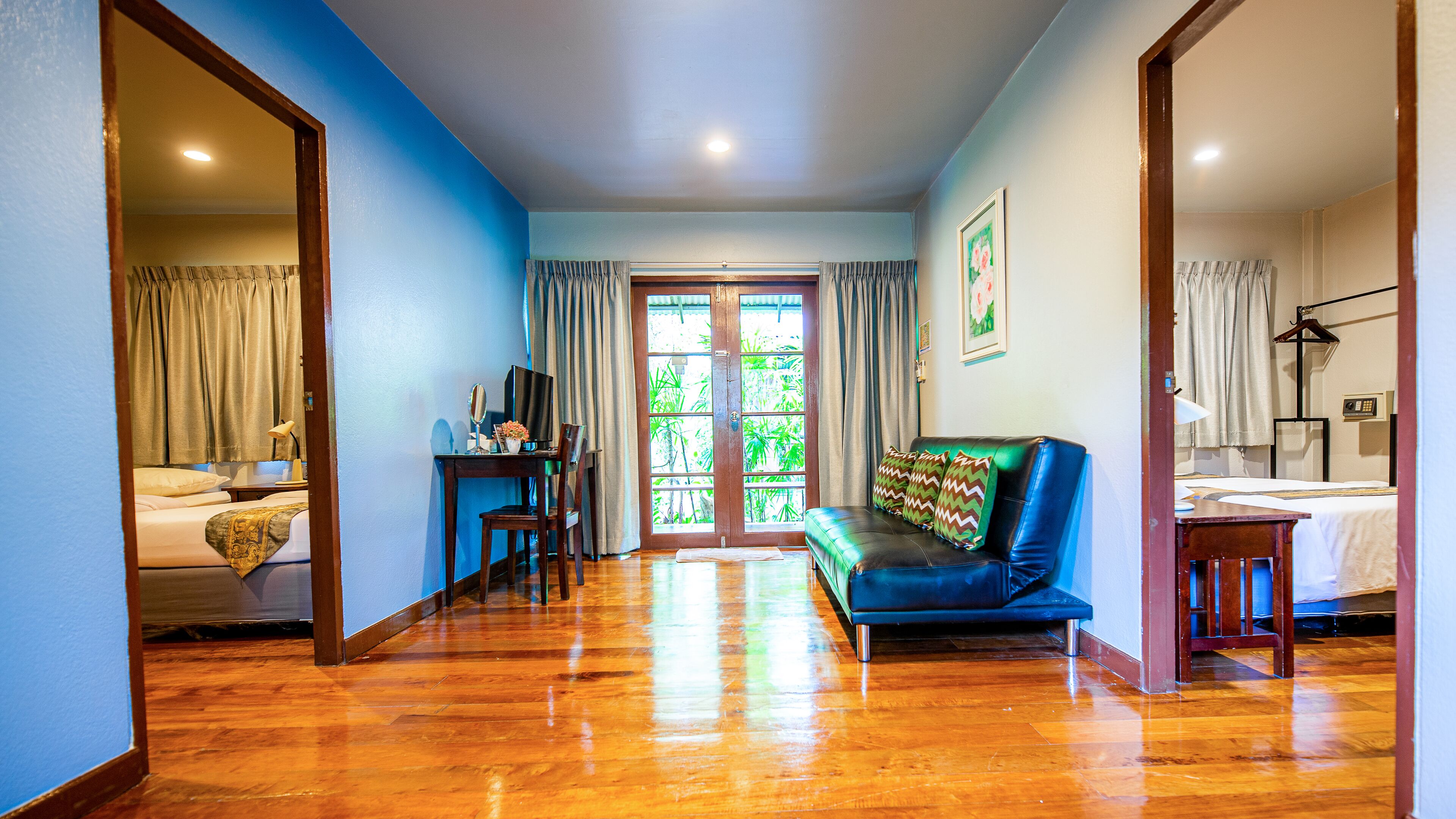 A hotel room with a wooden floor, a blue wall, a sofa with patterned cushions, a desk with a computer, and a bed with a patterned bedspread.