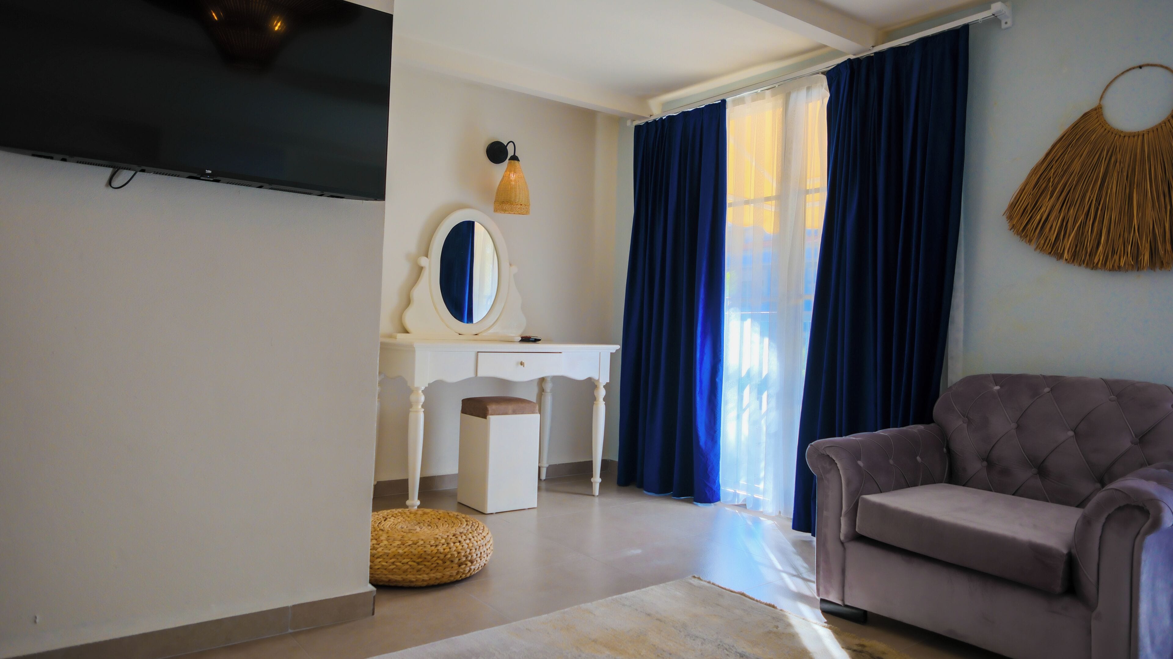 A room with a television, a white vanity with a mirror, a grey sofa, and blue curtains.