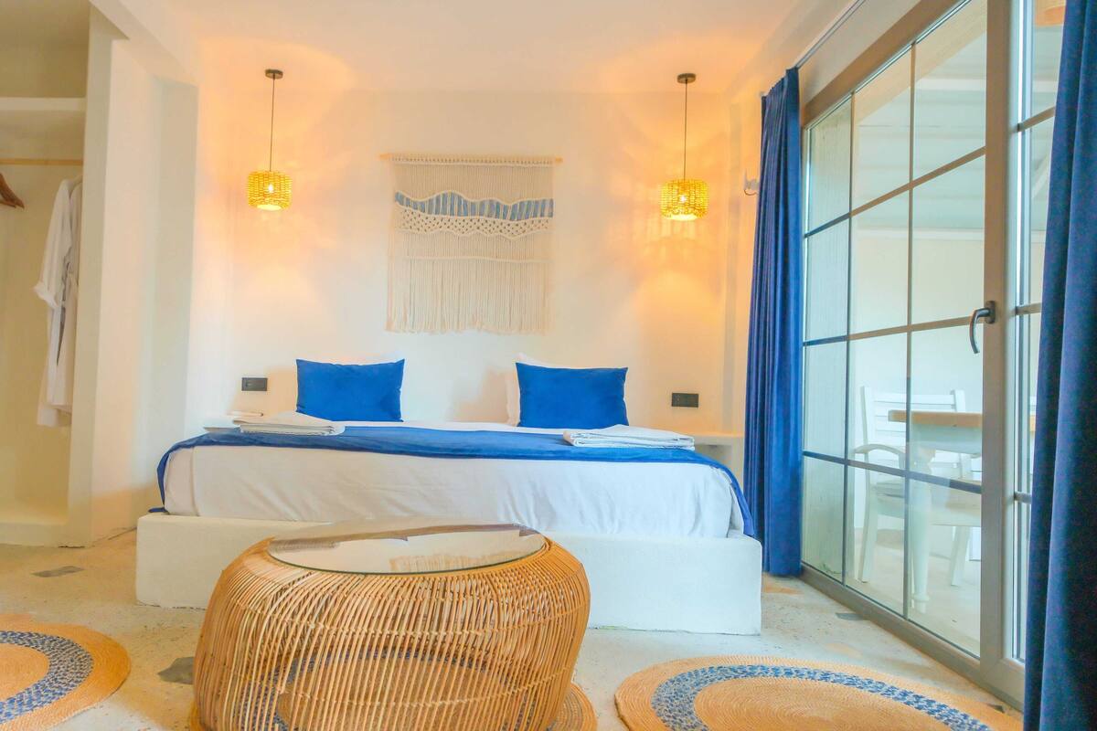 A bedroom with a bed, two wall-mounted lights, a woven chair, and a large window with curtains.