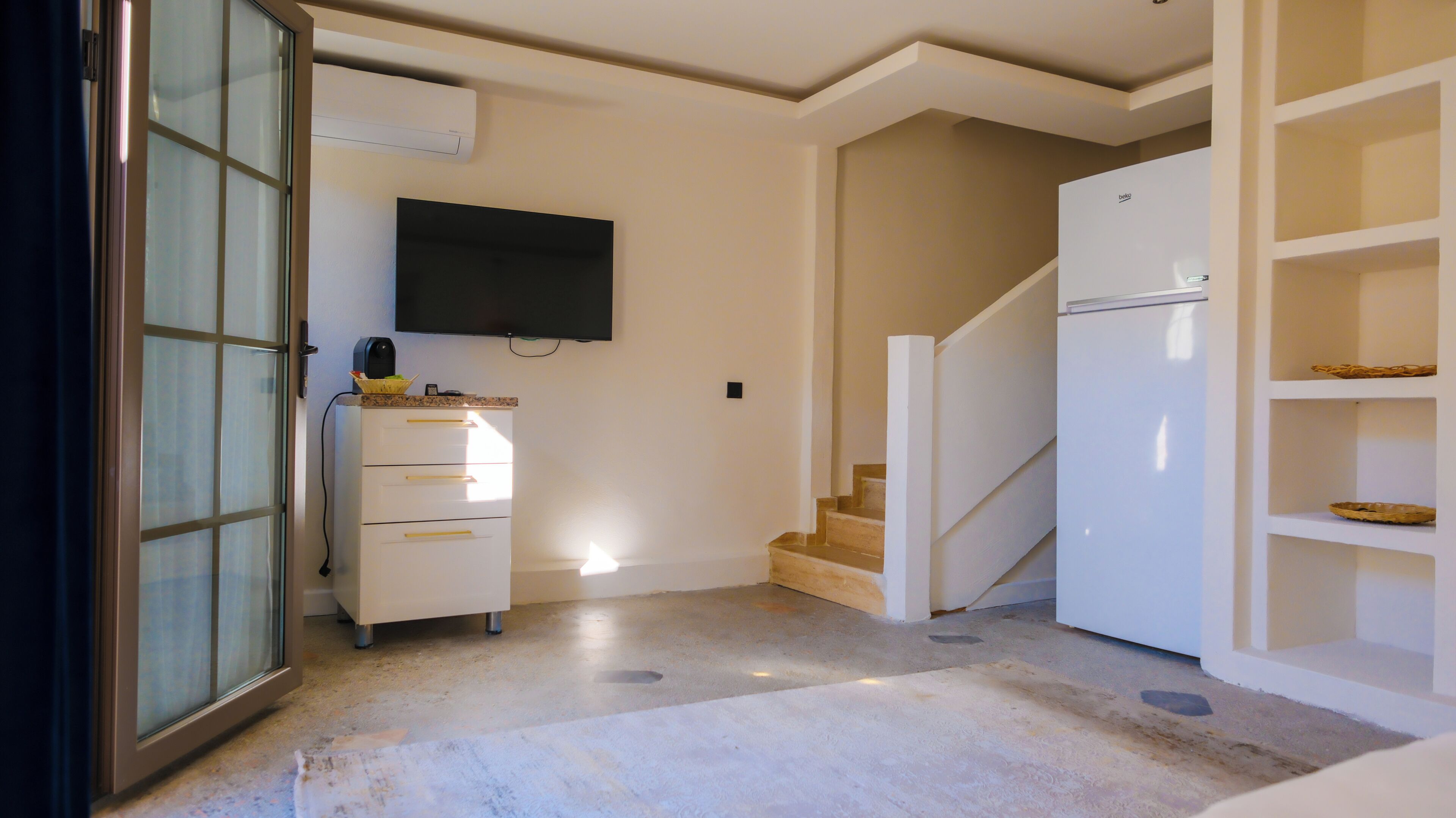 A hallway with a white refrigerator, a flat-screen TV mounted on the wall, a white cabinet with drawers, and a staircase leading to another level.