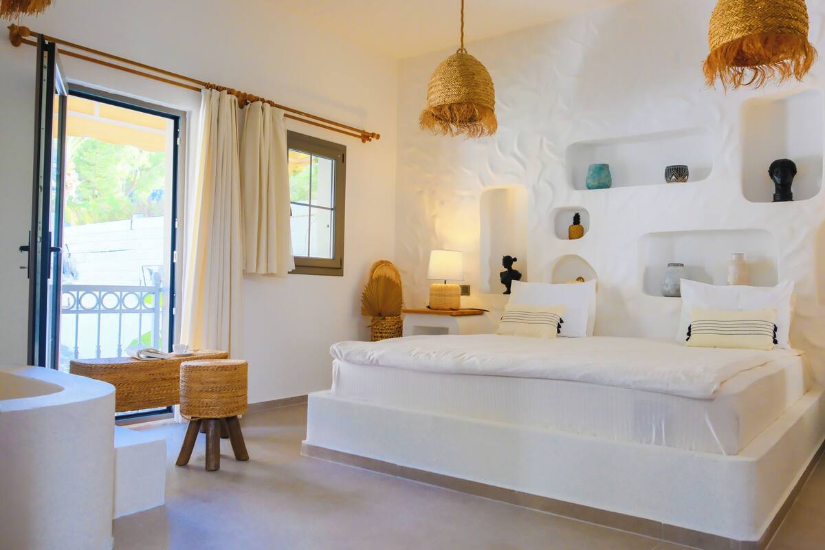 A white-themed bedroom with a bed, bedside tables, a lamp, a wicker stool, and a view of the outdoors through a door.