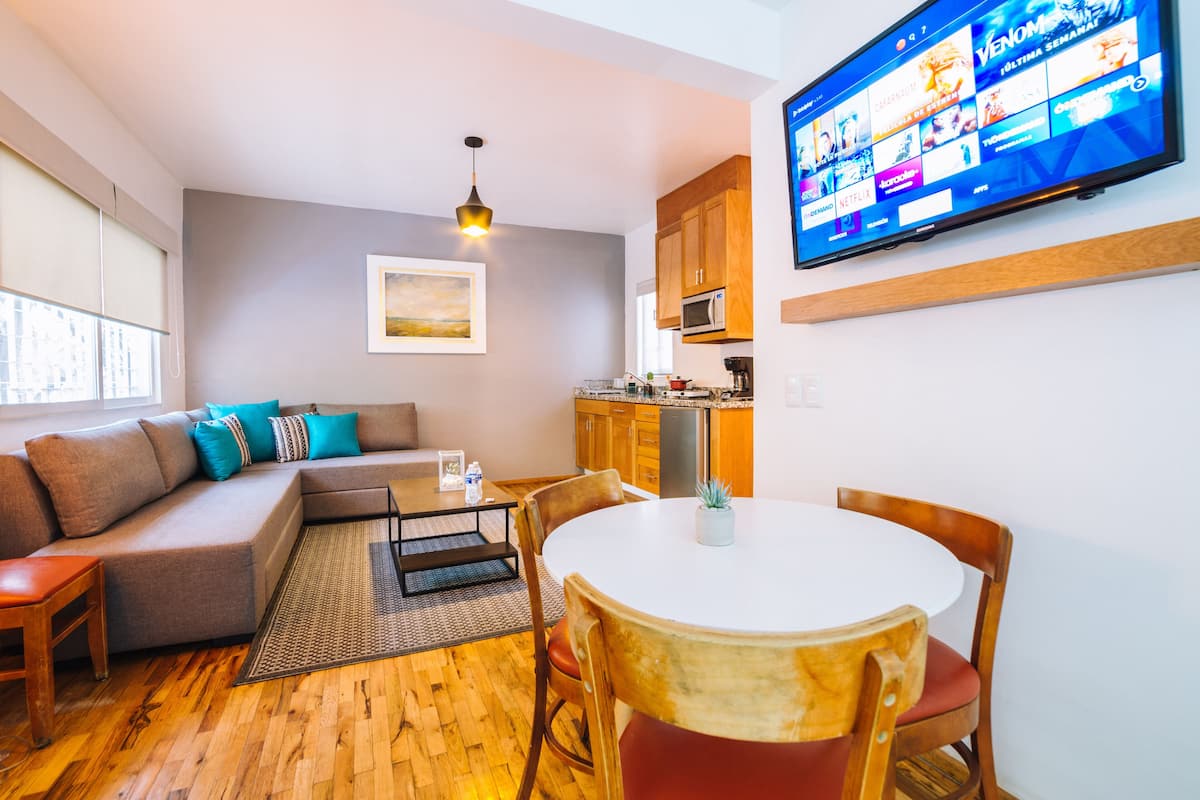 Ein modernes Wohnzimmer mit einer Couch, einem Esstisch mit Stühlen, einer Küchenzeile und einem an der Wand befestigten Flachbildfernseher.