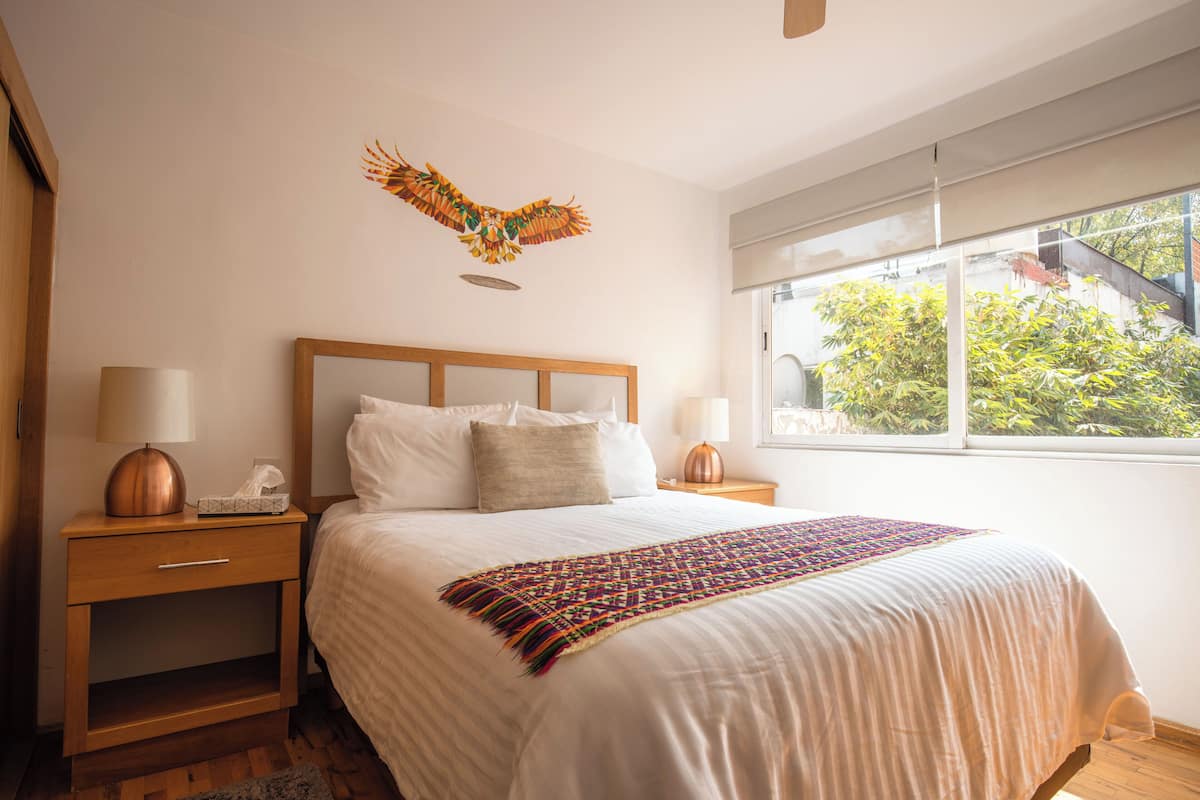 Ein ordentlich gemachtes Bett mit farbenfroher Tagesdecke, ein Nachttisch aus Holz mit einer Lampe und ein Fenster mit Blick auf Grünfläche.