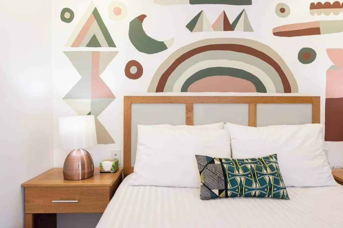 Ein ordentlich bezogenes Bett mit weißen Leinen und einem gemusterten Kissen, ein Nachttisch aus Holz mit einer Lampe sowie eine Wand mit abstrakter geometrischer Kunst.