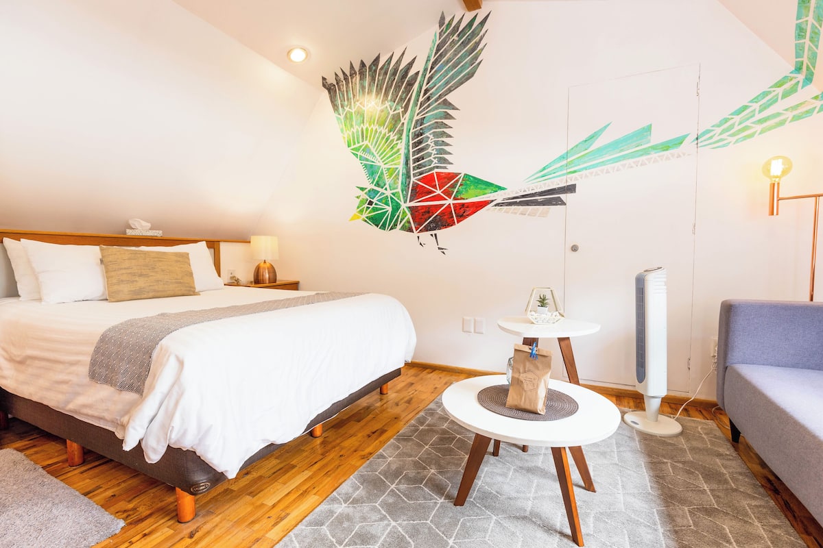 Ein modernes Hotelzimmer mit einem Bett, einem kleinen runden Tisch, einem Standventilator und einem Wandbild.