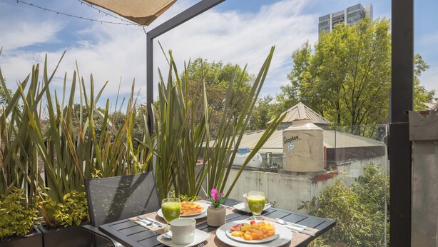 En balkon med et bord dækket til morgenmad, inklusive frugtfade og to glas grøn drik, omgivet af høje planter og udsigt til bygninger.