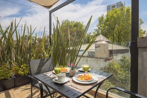 A balcony with a table set for breakfast, including plates of fruit and two glasses of green beverage, surrounded by tall plants and a view of buildings. - The Gallery by MyPlace (Mexico City)