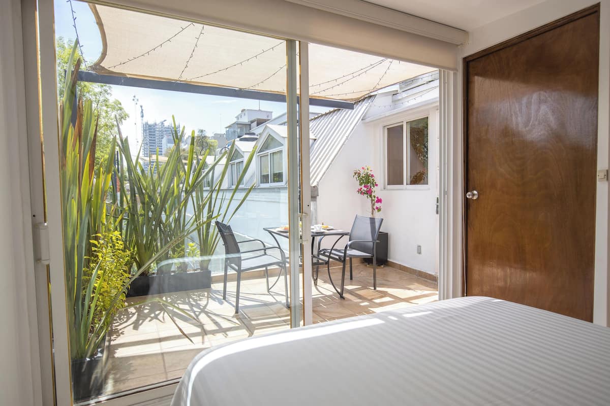 Ein Schlafzimmer mit einem großen Bett, einer Schiebetür zu einem Balkon mit Tisch und Stühlen und Blick auf die Stadtlandschaft.