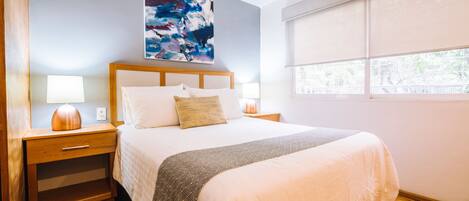 A modern bedroom with a large bed, wooden headboard, bedside tables with lamps, a nightstand, a rug, and a window with blinds.