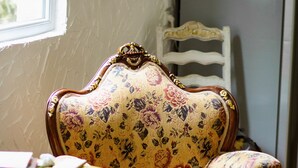 Ein alter, floral gemusterter Sessel mit aufwendigen Holzbeinen, neben einem runden Tisch mit gelber Tischplatte.