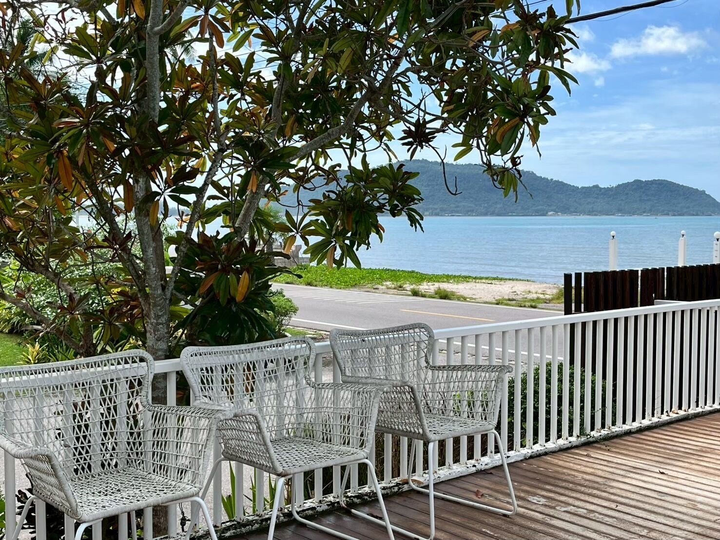 Una terraza de madera con sillas metálicas blancas y una cerca blanca, con vistas a un cuerpo de agua y colinas distantes.