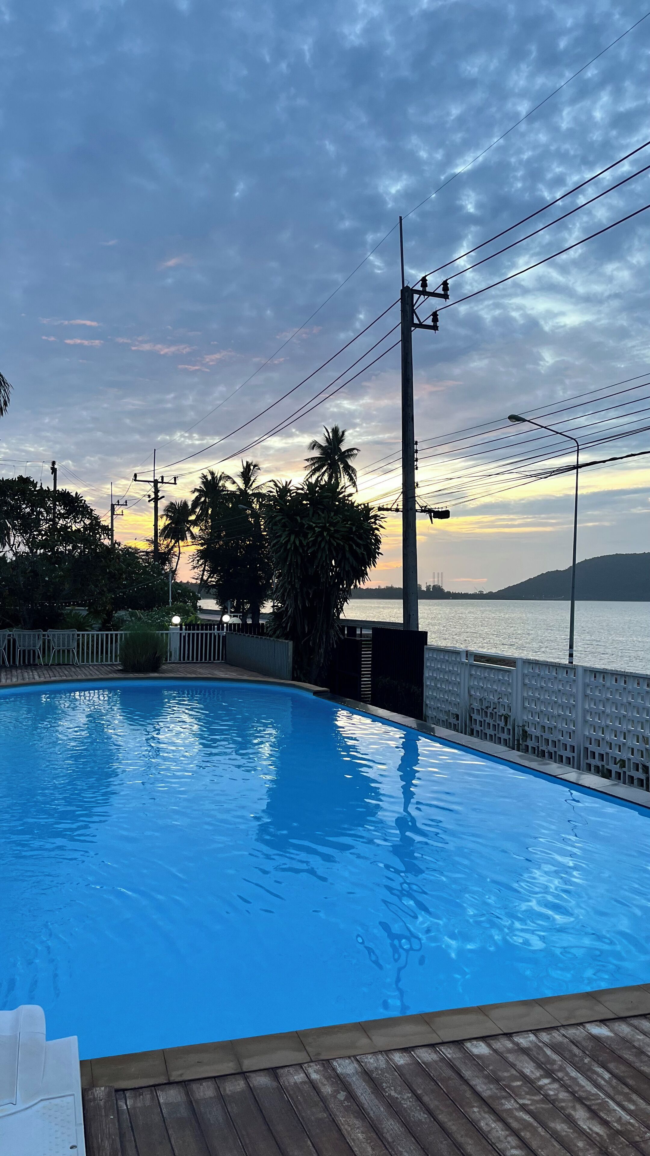 Una piscina con cubierta de madera, rodeada por una cerca y palmeras, con vista a una masa de agua y un atardecer al fondo.