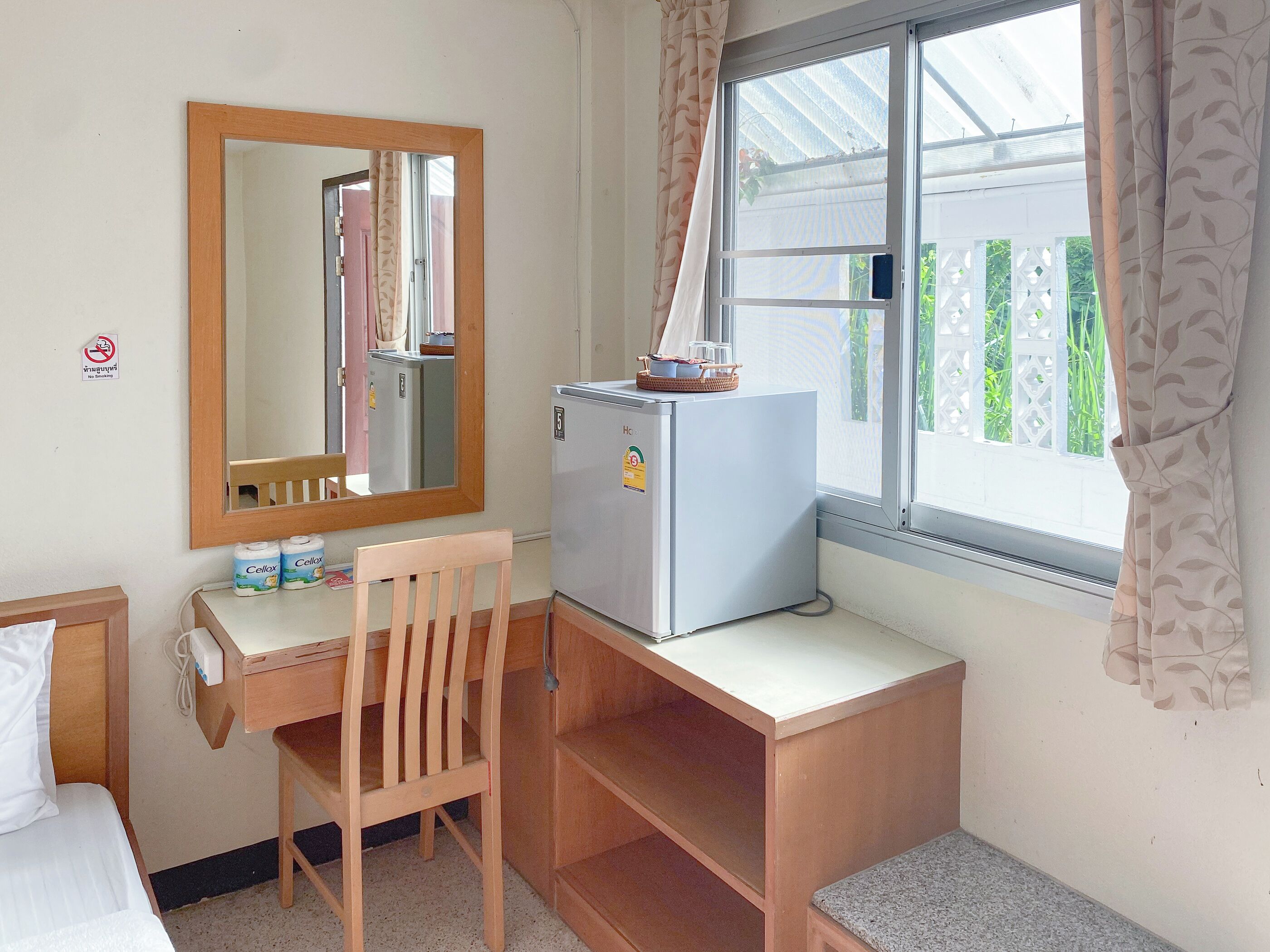 Een compacte hotelkamer met een eenpersoonsbed, een houten bureau, een kleine koelkast, een stoel, een spiegel en een raam met gordijnen.