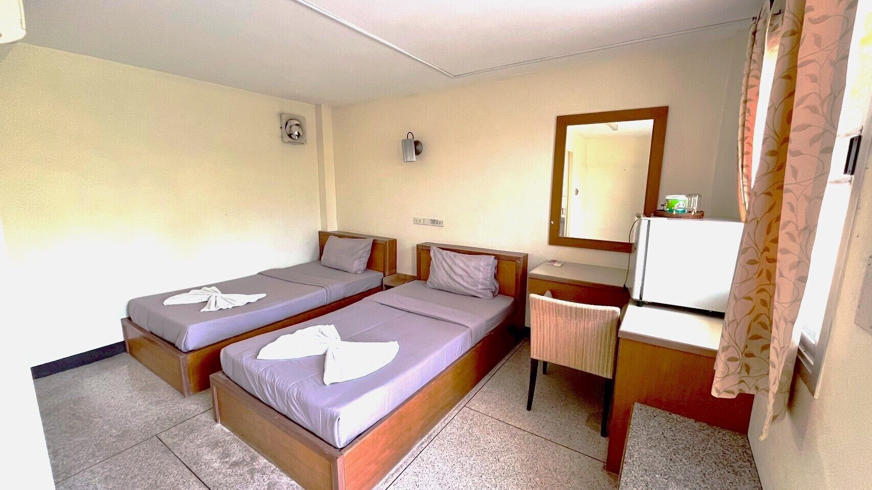 Una habitación de hotel con dos camas individuales, un escritorio con silla, un espejo y un ventanal con cortinas.