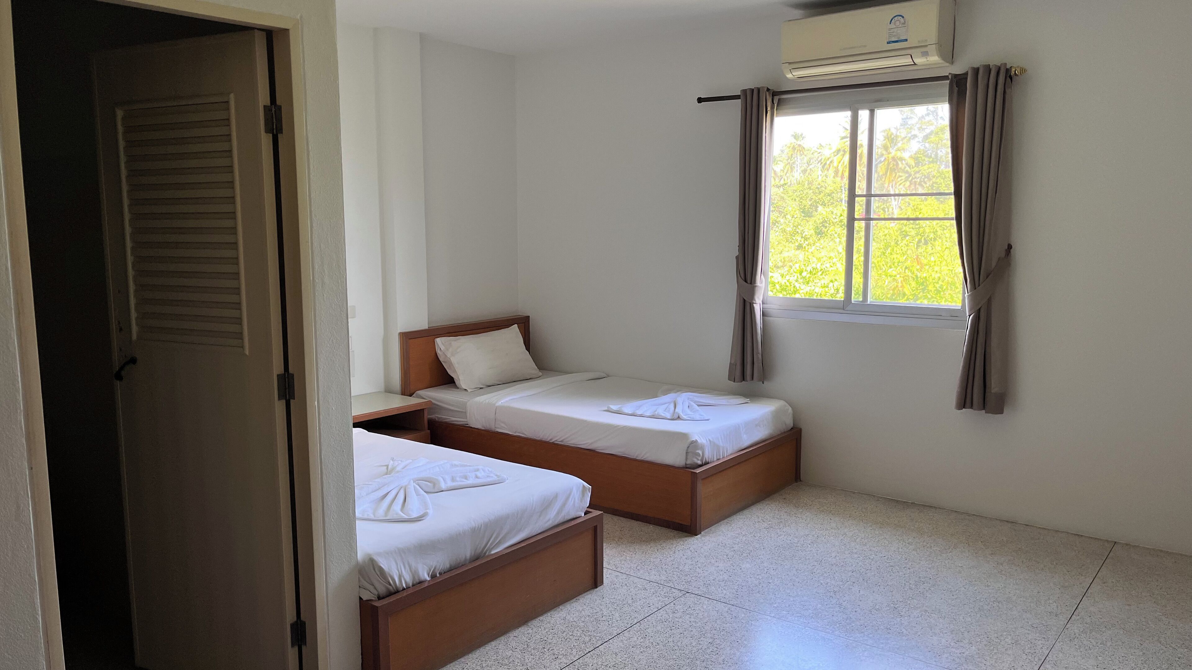 A room with two single beds, a window with curtains, a door, and an air conditioning unit.