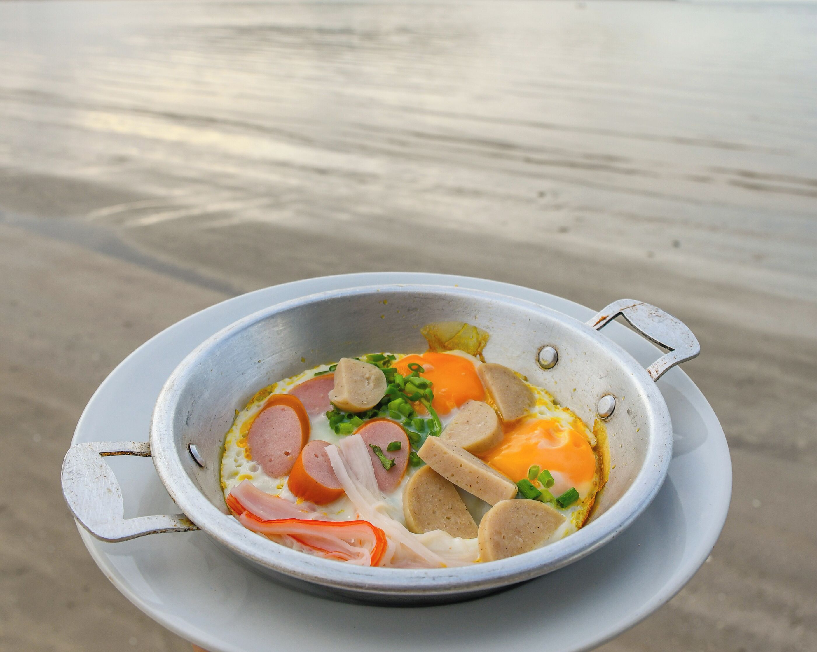 A metal bowl with a white interior containing sliced sausage, eggs, and green onions, placed on a white plate.