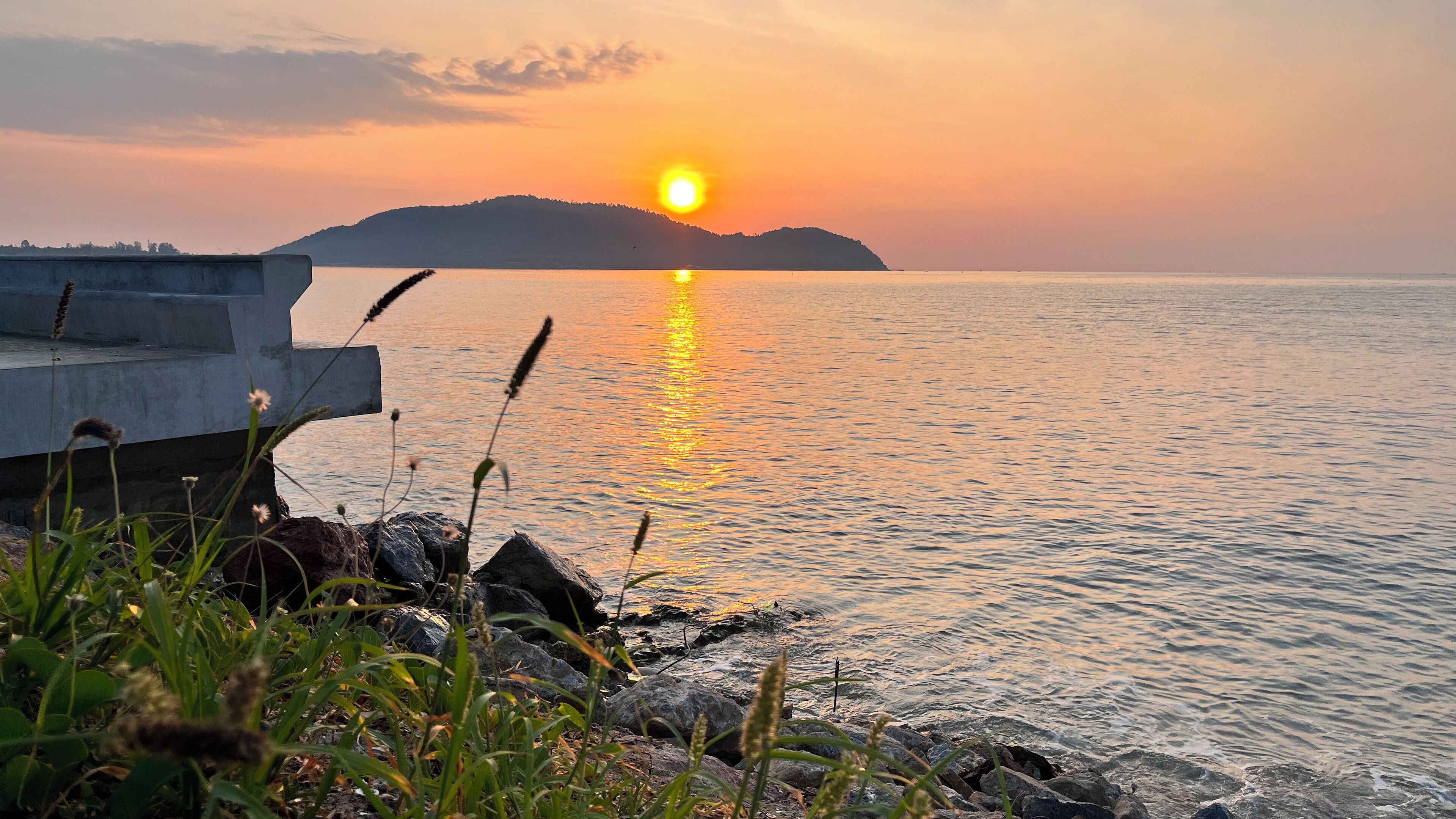 A sunset over a calm body of water with a rocky shoreline and a distant hill.