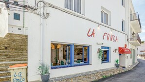 A white building with the sign "LA FONDA HOSPITAL" indicating a hospital. There are windows with blue frames, a red awning, and a stone wall with a trash bin labeled "La Fonda HOSPITAL". - Hostal La Fonda (Cadaqués)