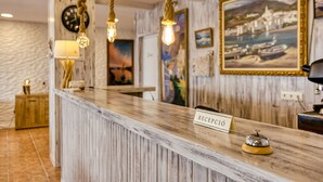 A reception desk with a 'RECEPCIÓN' sign, rustic wooden paneling, a clock, and a framed painting of a coastal scene. - Hostal La Fonda (Cadaqués)