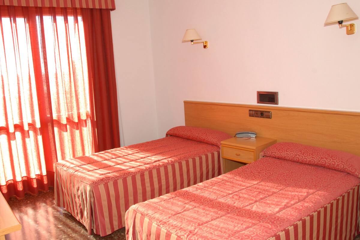 A hotel room with two single beds, a nightstand, a lamp, and a window with curtains.