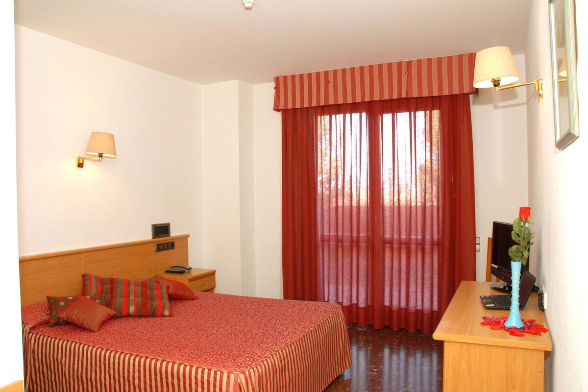 A hotel room with a bed, bedside table, lamp, and a desk with a computer and vase.