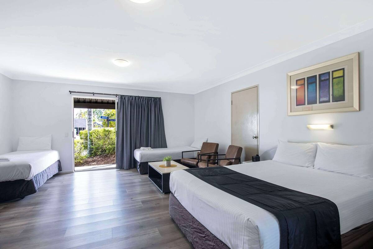 A hotel room with two single beds, a large bed, a desk with a chair, and a view of the outdoors.