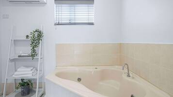 Ein Badezimmer mit einer Badewanne, Ablagefläche mit gefalteten Handtüchern, einer Topfpflanze und einem Fenster mit Jalousien.