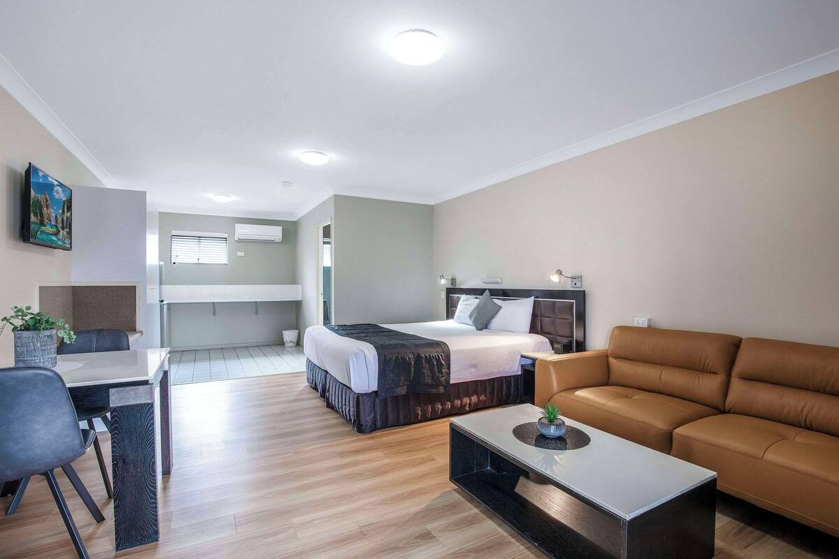 A modern hotel room with a bed, a sofa, a dining table, and a small kitchen area.