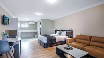 A modern hotel room with a bed, a sofa, a dining table, and a small kitchen area.