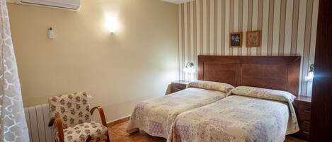 A bedroom with two beds, a chair, a table, an air conditioner, and a wall-mounted light fixture.