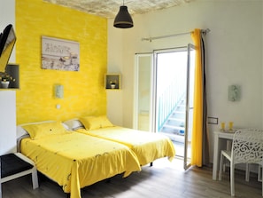 Una habitación con dos camas cubiertas con ropa de cama amarilla, una mesita con dos vasos, una silla blanca y vistas a una escalera.