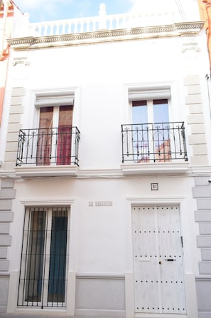 Un edificio blanco con dos balcones, una ventana con rejas y una puerta con placa de número.