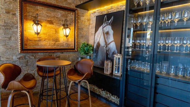 A bar area with a stone wall, two wall-mounted lights, a large framed horse portrait, and a glass display case with wine glasses. - Brii hotel (Araguaina)