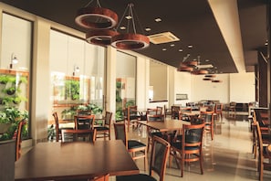 A spacious dining area with wooden tables and chairs, large windows with plants, and modern pendant lighting. - Brii hotel (Araguaina)