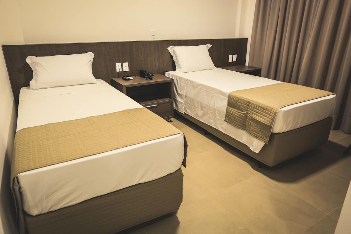 A hotel room with two single beds, each with a pillow and a blanket. There is a bedside table with a phone and a remote control.