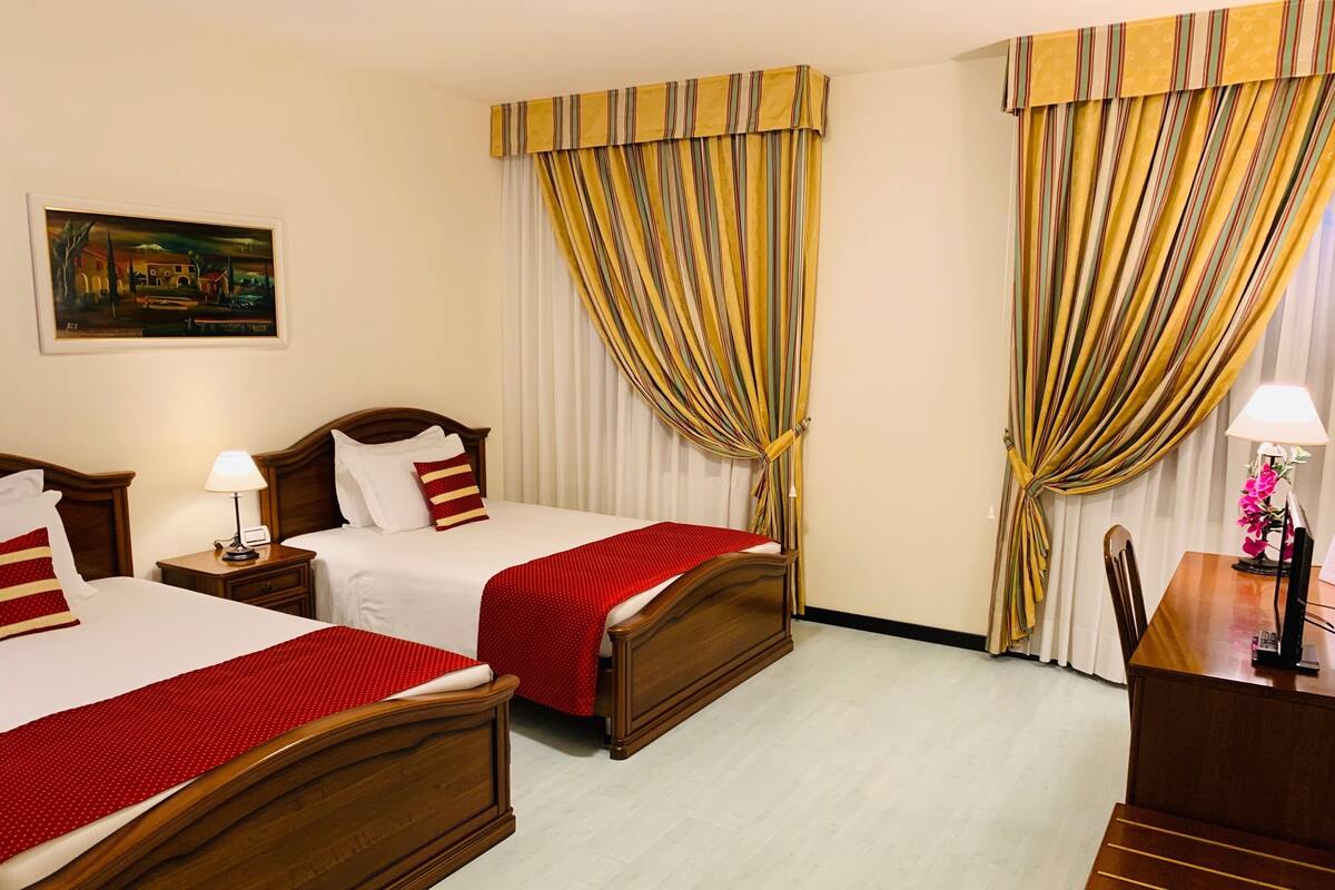 A hotel room with two wooden beds, a desk, a chair, a lamp, and curtains.