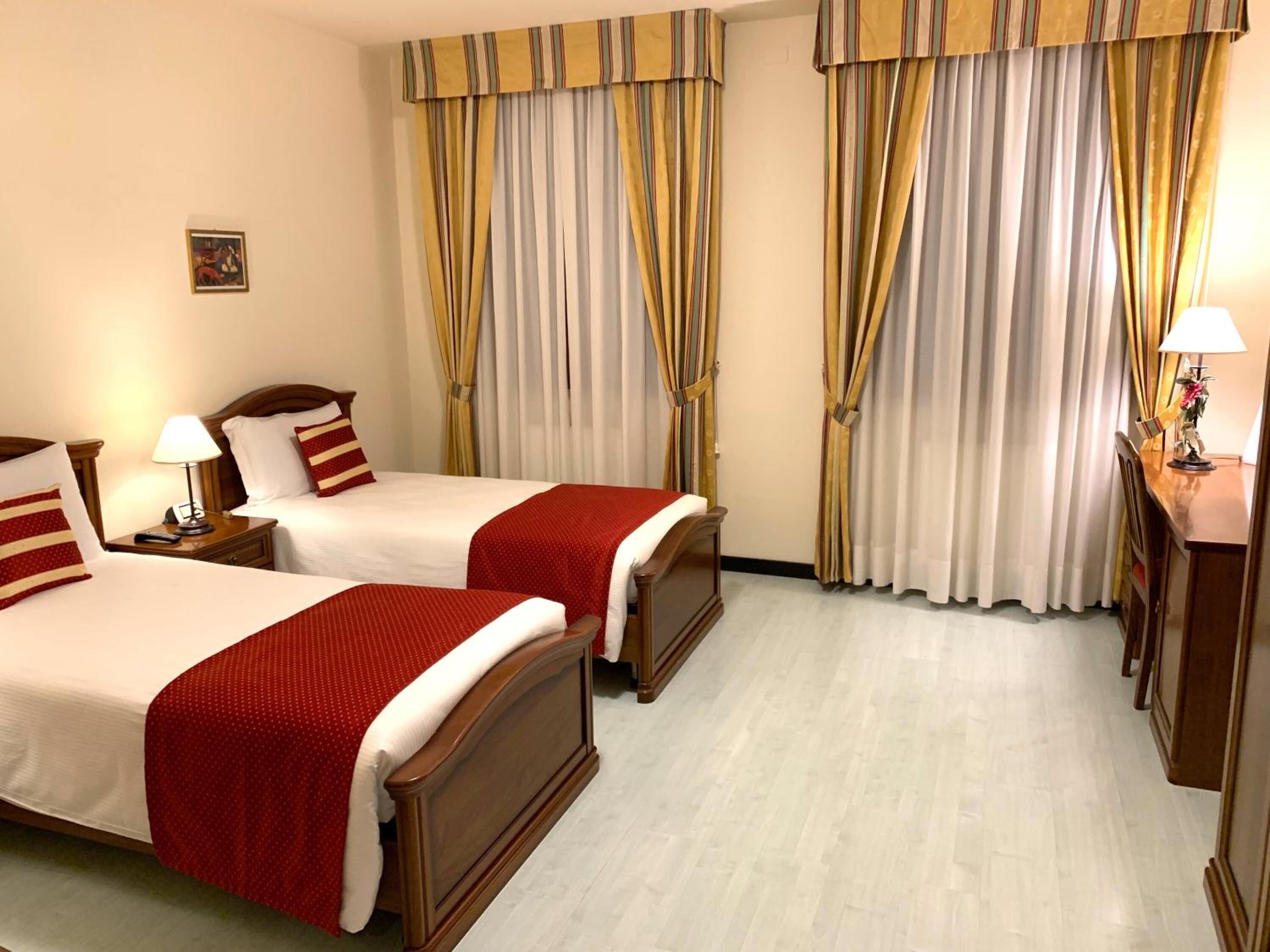 Une chambre d’hôtel avec deux lits, chacun doté d’une tête de lit en bois, de draps blancs et d’éléments décoratifs rouges. La chambre a des murs de couleur claire, des rideaux à rayures et un bureau en bois avec une lampe.