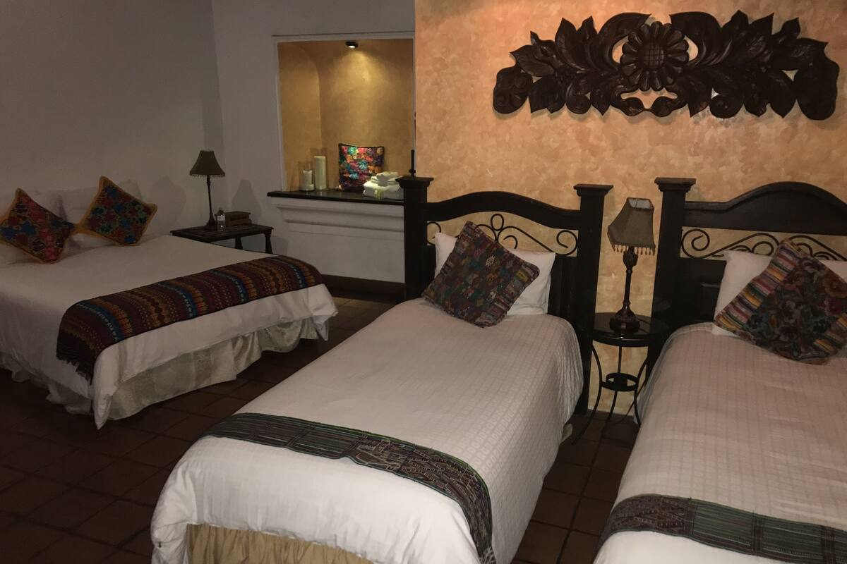 Habitación de hotel con dos camas, cada una con una colcha estampada y múltiples almohadas. Hay una mesita de noche con una lámpara y una pequeña mesa con un adorno. La habitación tiene un ambiente cálido y acogedor con un objeto decorativo sobre las camas.