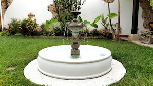 Une fontaine ronde, dotée d’un piédestal en pierre et dont l’eau jaillit du sommet, est entourée d’une pelouse bien entretenue et de diverses plantes.